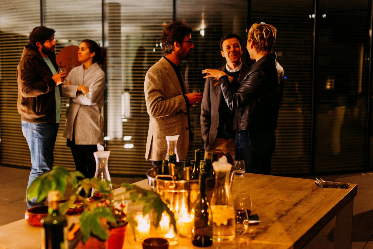 An elegant evening social gathering in a modern kitchen or dining area. Five people are mingling and conversing, dressed in smart casual attire including blazers and leather jackets. In the foreground, a wooden table displays an array of wine bottles, glasses, candles, and a potted plant, creating an intimate dinner party atmosphere. The lighting is warm and ambient, with horizontal blinds visible in the background and modern fixtures creating a sophisticated setting for the social event.